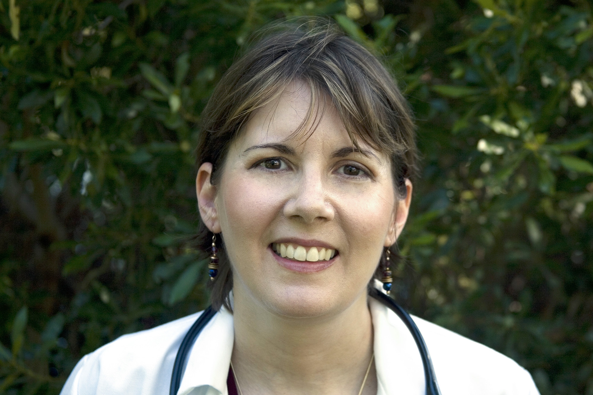 Amazon author CJ Lyons smiles into the camera. She has dark hair and is wearing a white jacket with a stethoscope around her neck. The background is blurred trees. 