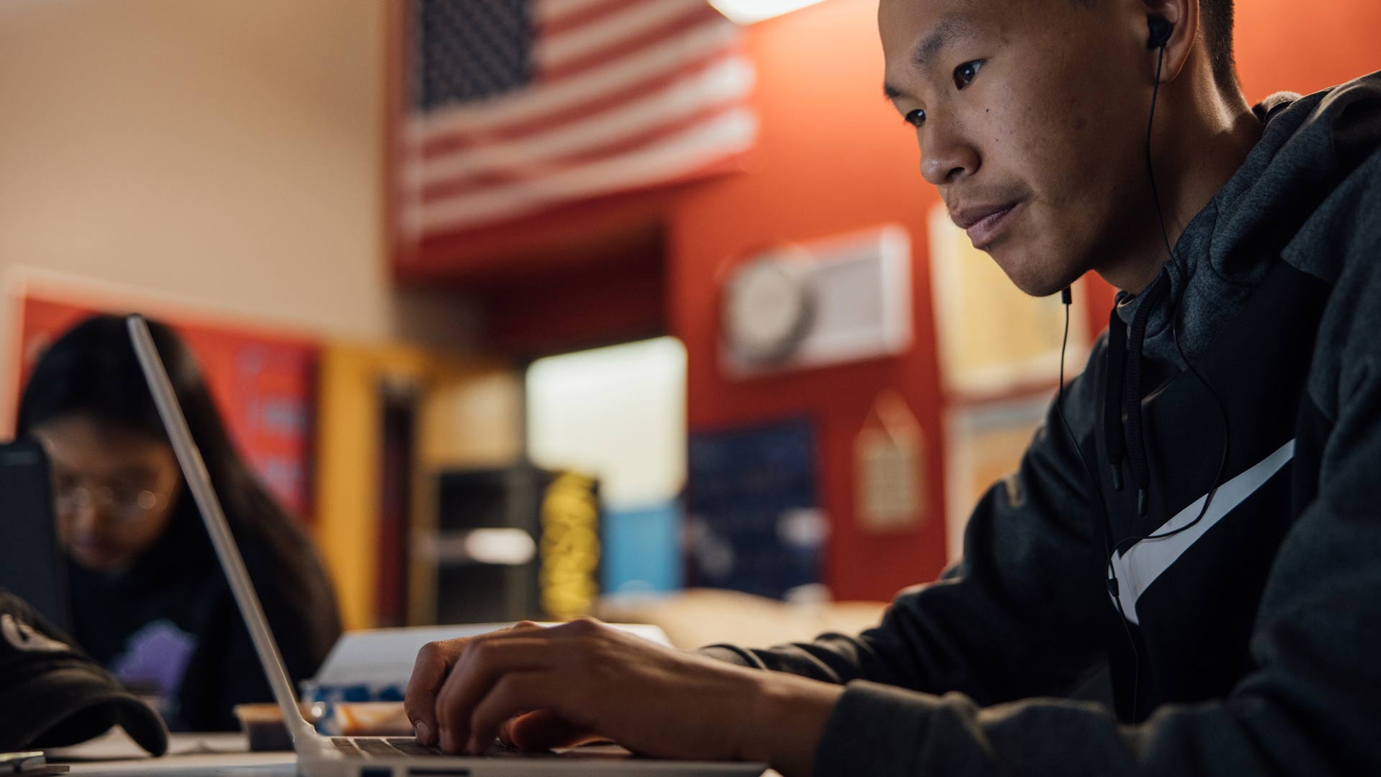 A student works on a laptop with the U.S. flag in the background.
