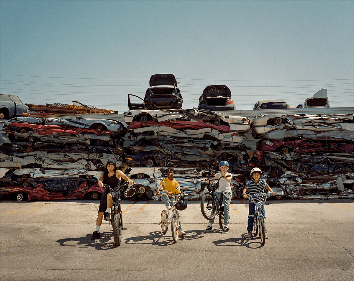 An image of four kids standing on their bikes while looking at the camera. Behind them is a junkyard of smashed cars.