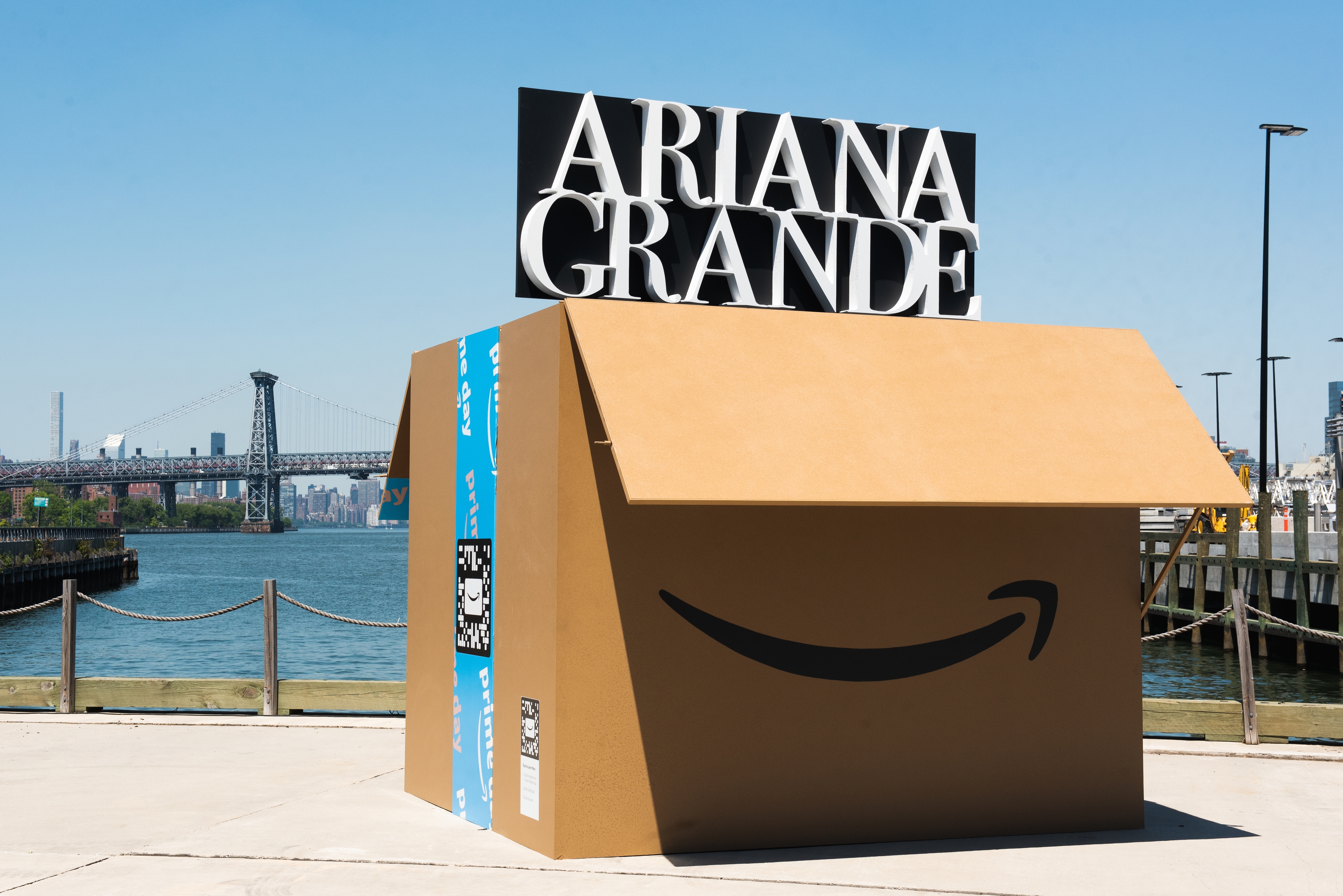 A large Amazon box on the waterfront. The box is opened on top, to reveal the name of musical artist, Ariana Grande, in white letters on black background. 