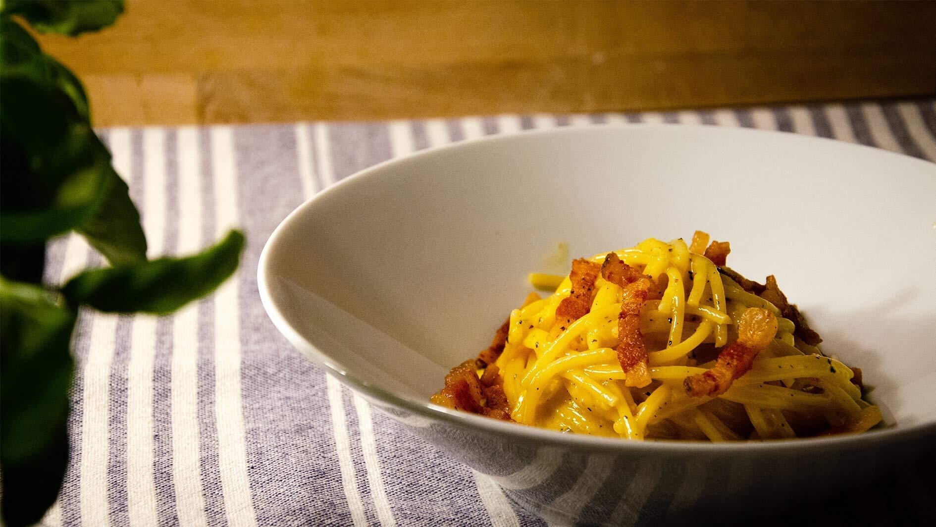 An image of a bowl sitting on a blue and white striped table cloth. The bowl has pasta with bacon inside of it.