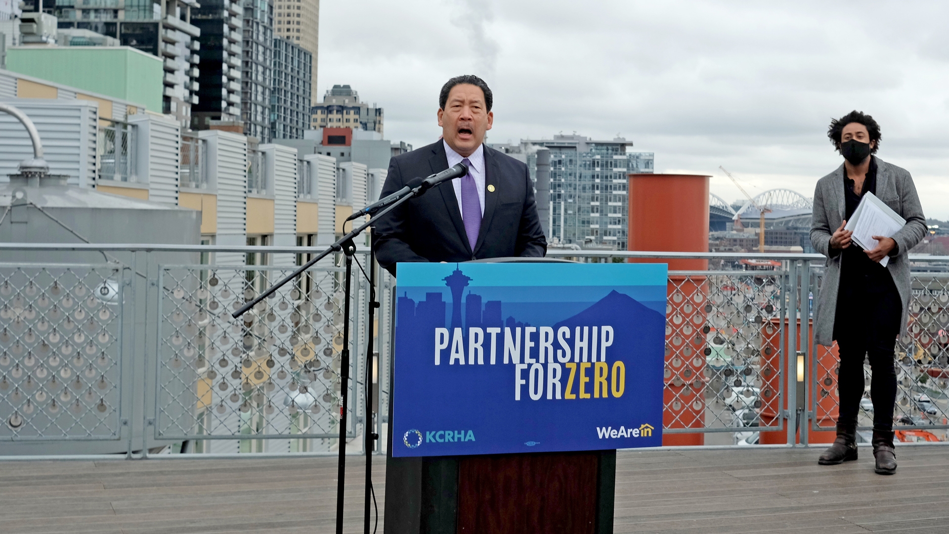 The Mayor of Seattle, Bruce Harrell, speaks into a microphone as he stands behind a podium with a sign that reads, "Partnership for Zero."