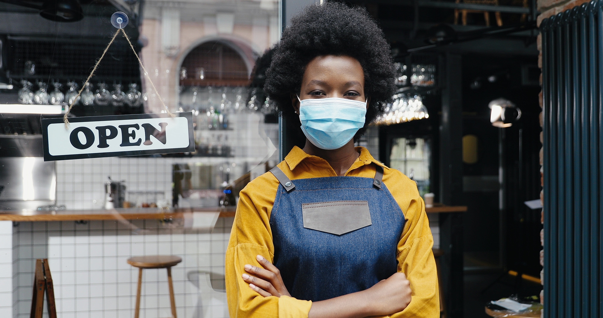 A Black woman wearing a mask and a denim apron stands in the open doorway of a restaurant