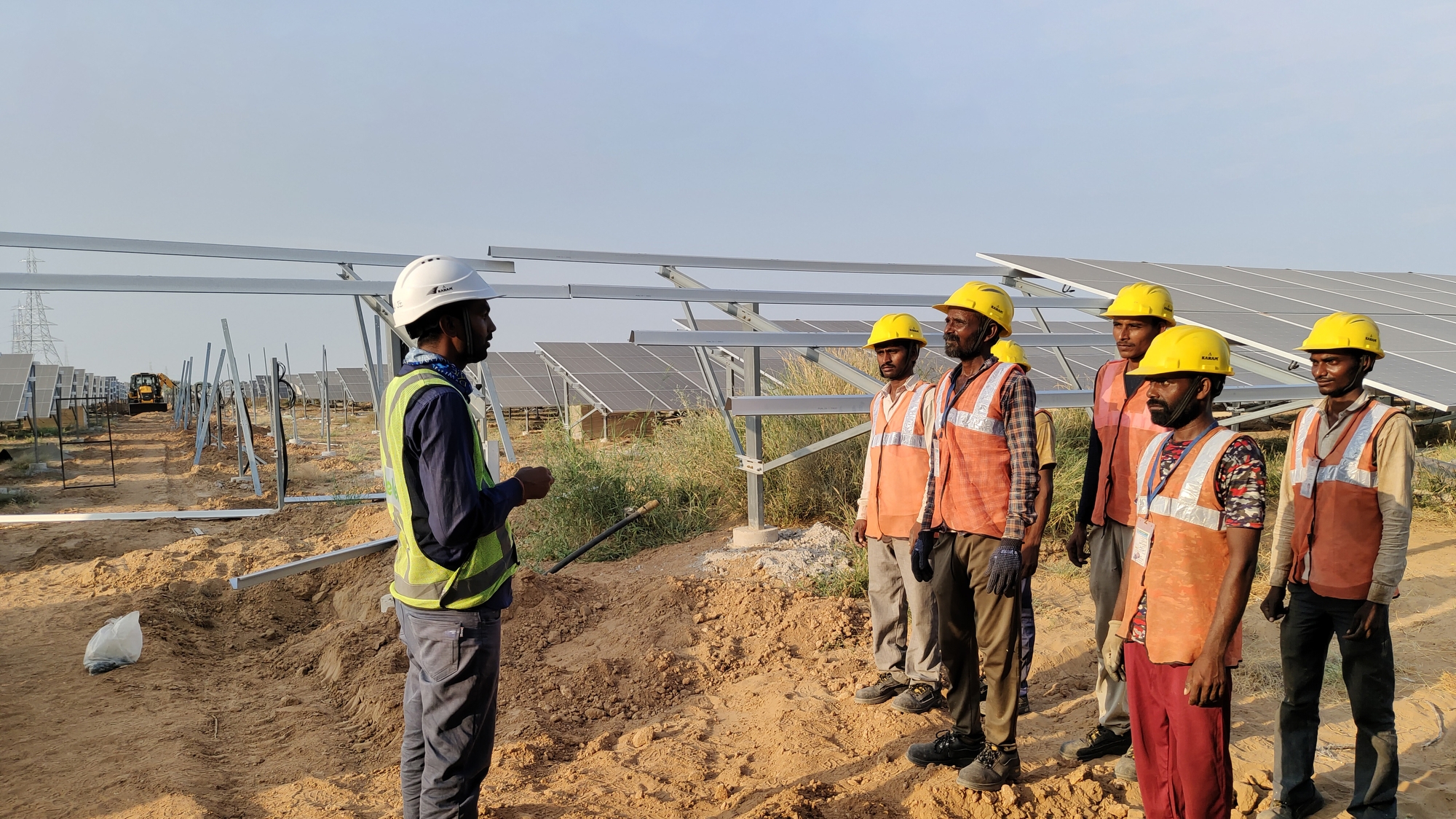 An image of a group of employees in India having a conversation in a solar farm. They are all dressed in vests and helmets.