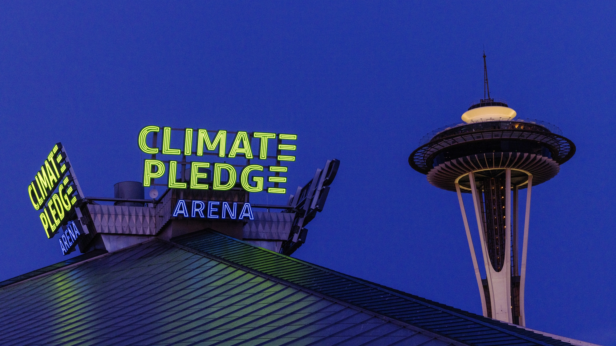 An image of the top of Climate Pledge Arena in Seattle at night. There is the illuminated neon sign that says "Climate Pledge Arena" on top of the stadium and the Space Needle in the background.