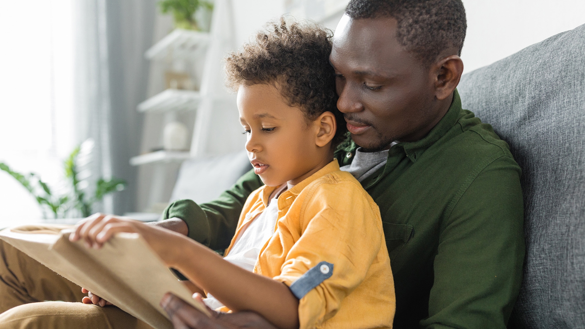 An image of a father and son sitting on a couch together. They are holding a book and the child is reading to his father.