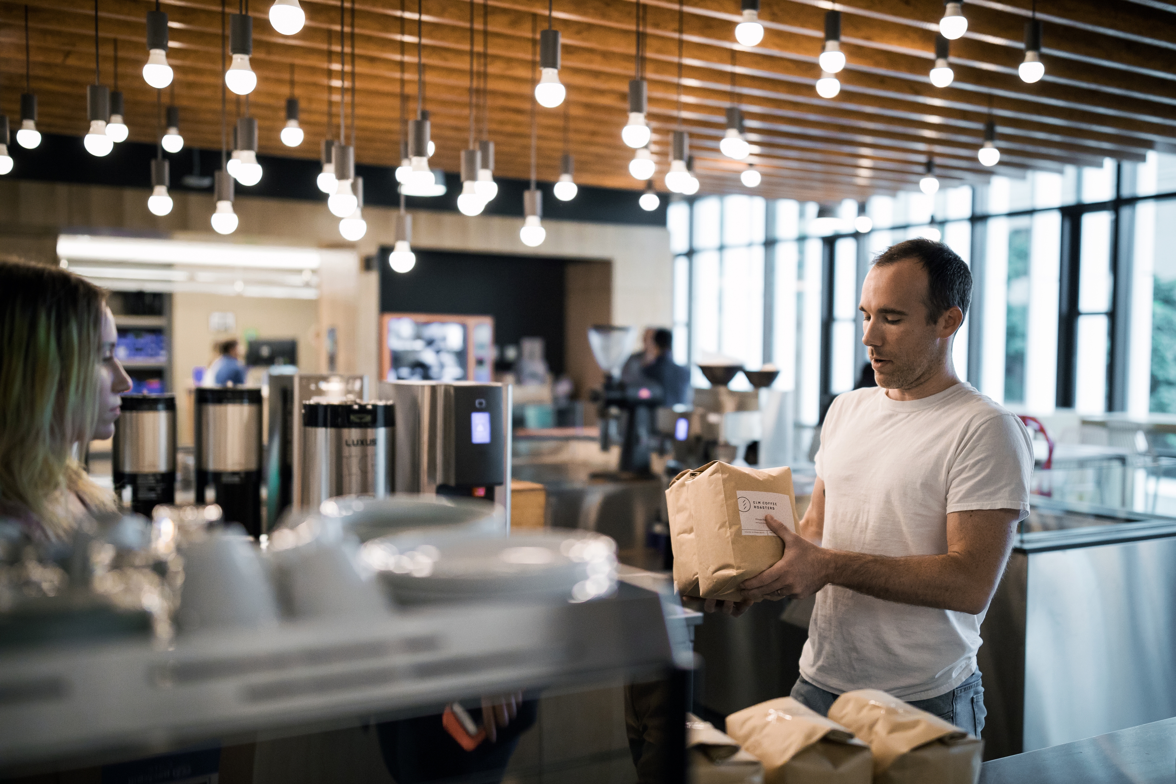 A male barista in a white t-shirt carries two bags of coffee towards a female customer. 