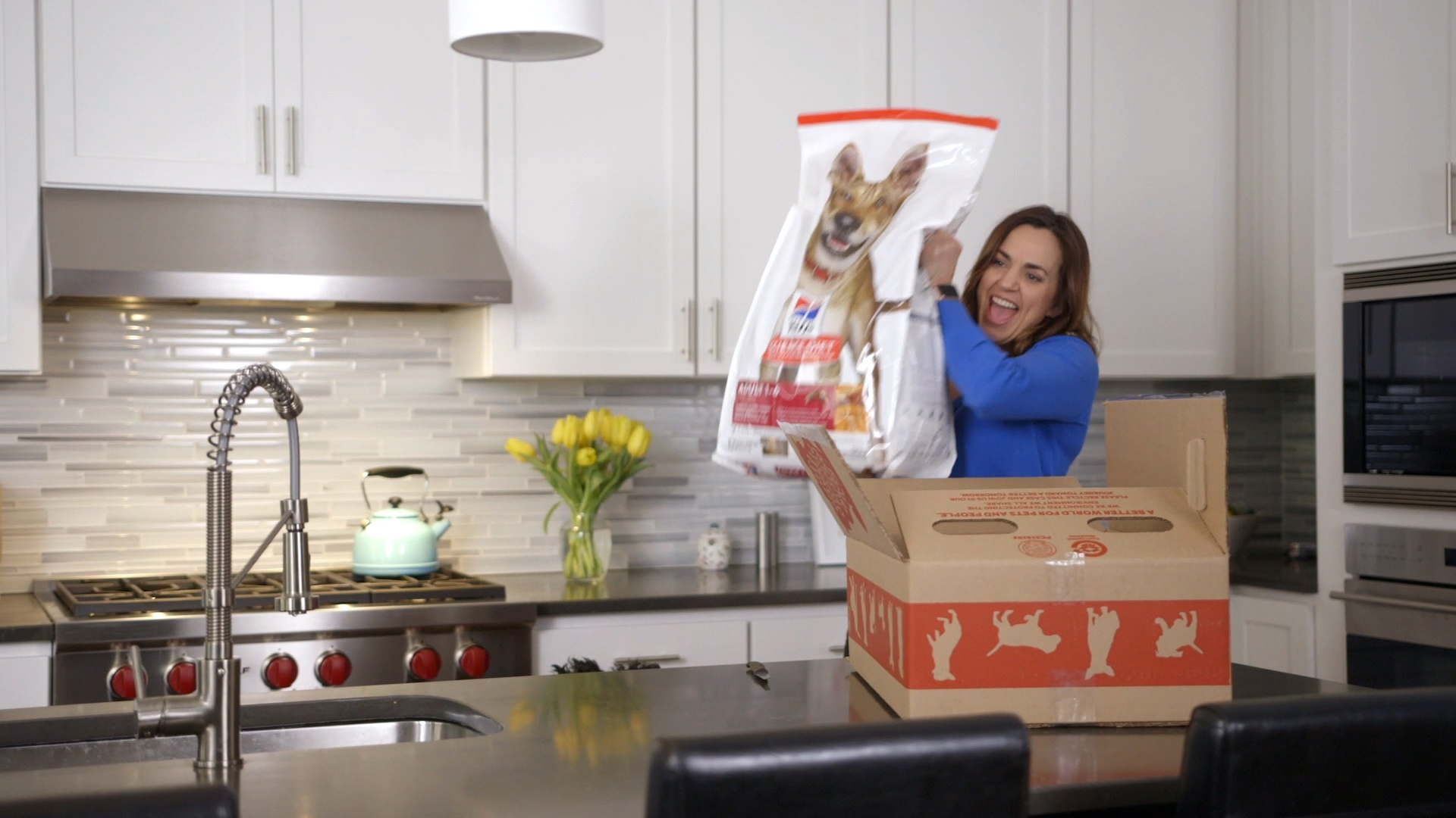 A woman lifts a bag of dog food from a cardboard box. She is standing in a kitchen.