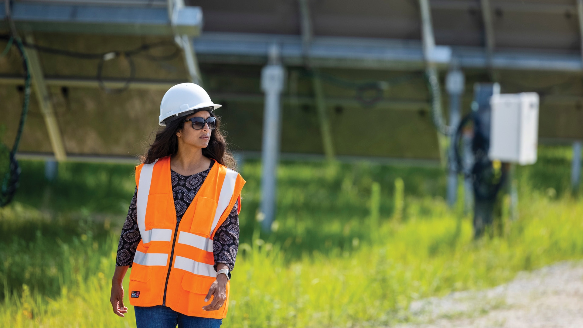 A woman wears an orange safey vest, a hard hat, and sungalsses as she walks outside by a solar farm.