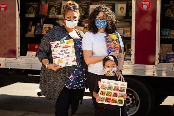 An image of two women standing with a child in front of a truck with books inside of it. They are each holding a book as they stand for the photo wearing face masks.