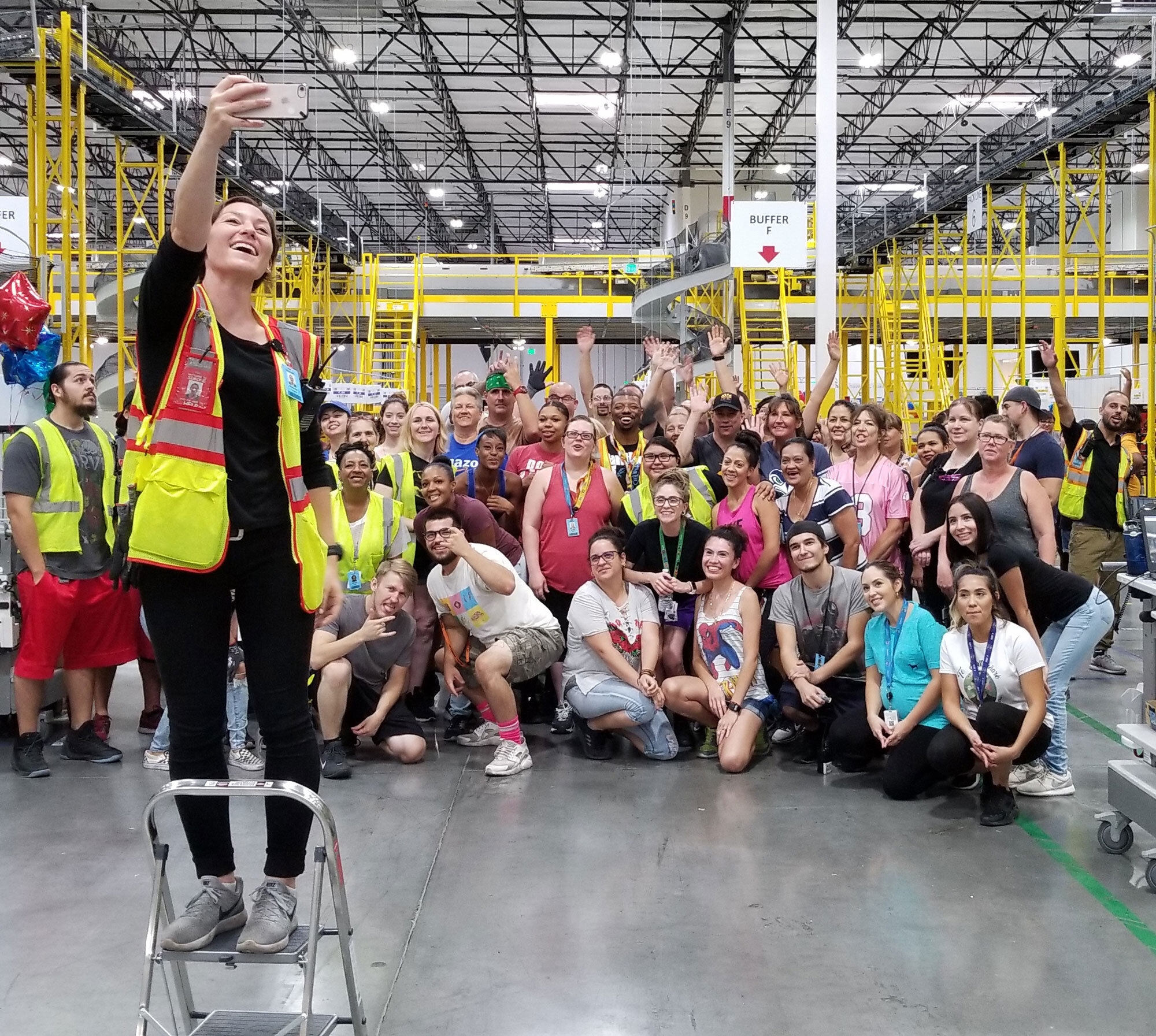 An Amazon associate stands on a step stool, with her arm extended, holding her iPhone in order to capture a selfie. Behind her, dozens of Amazon associates pose for the photo.