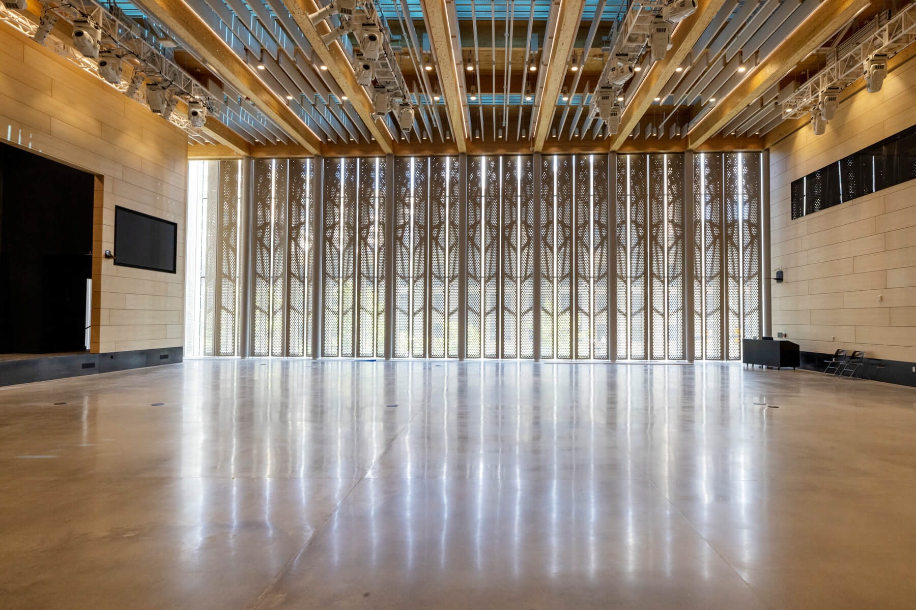 An image of a large event space at Amazon's second headquarters. It is a large room with a big window with elaborate shades and a ceiling full of lights with a large LED screen on one wall.