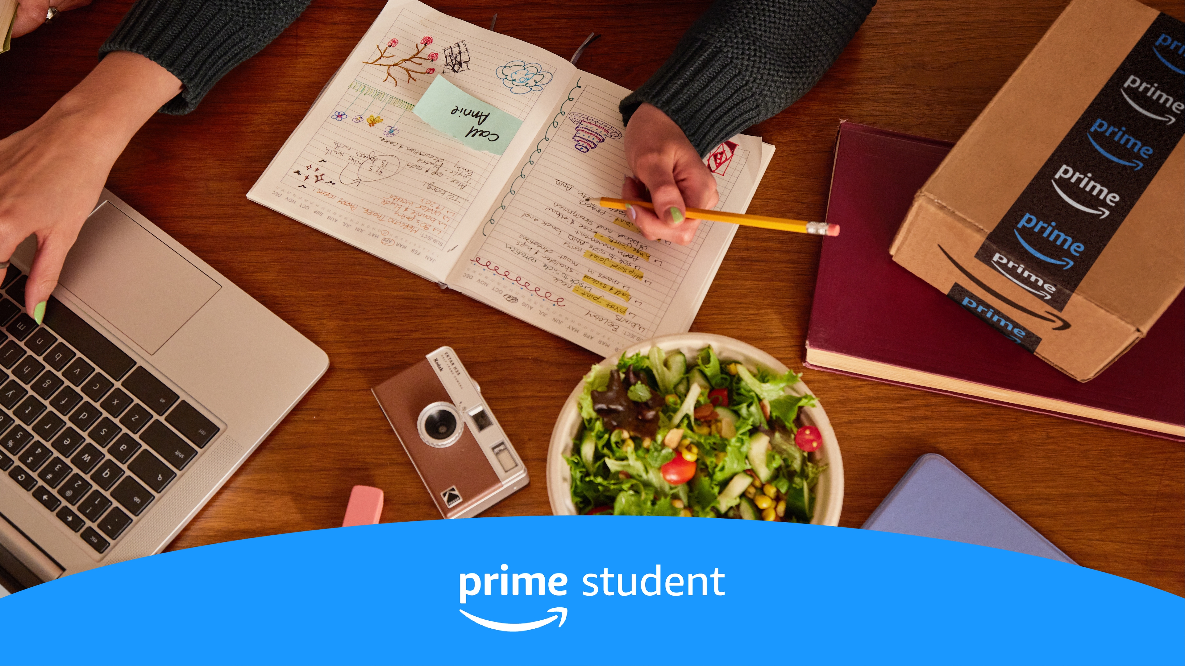 Image of a student studying with her notebook, camera, salad, and Amazon Prime delivery box on her desk.