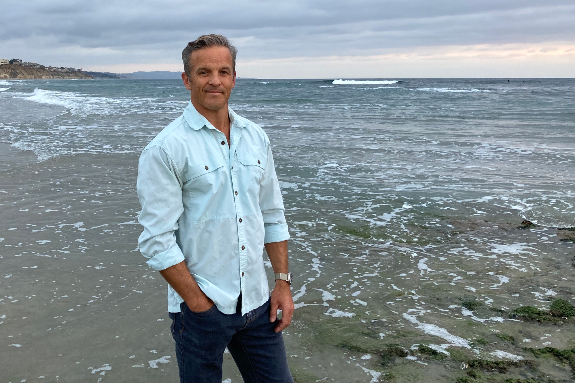 Author Dan Sheehan stands on an ocean beach wearing jeans and a button-down shirt.