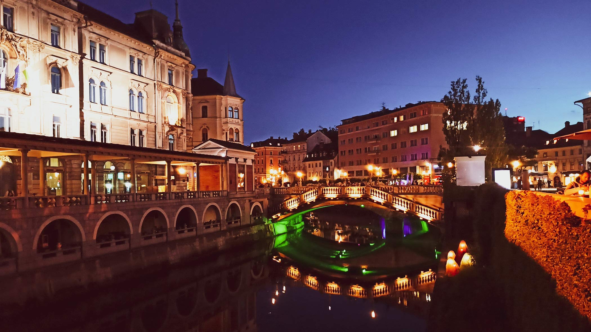 An image of a Slovenian city at night. There is a bridge over a canal with lights illuminating the bridge and surrounding buildings.