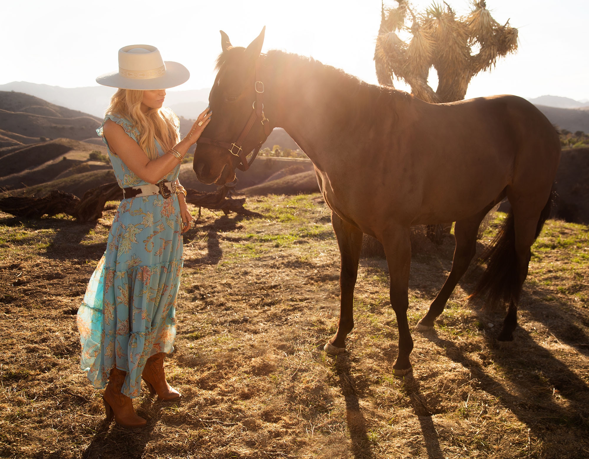 Jessica Simpson (singer, actress, author) stands in a field wearing cowgirl boots, a flowy dress, and belt, as she pets a horse on the muzzle in a promotional image
