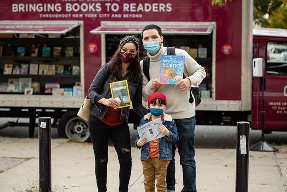 An image of a man and a woman standing with a child in front of a truck with books inside of it that reads "Bringing Books to Readers." All three people in the photo are holding up a book as they stand for the photo wearing face masks. 