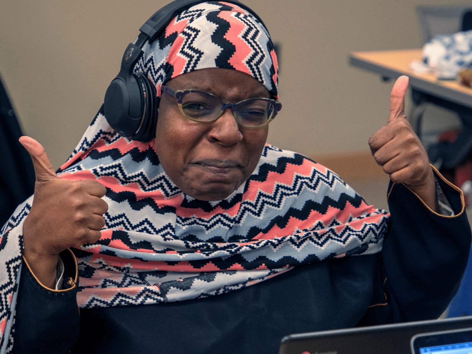 A photo of a woman wearing headphones, sitting in front of a computer holding two thumbs up.