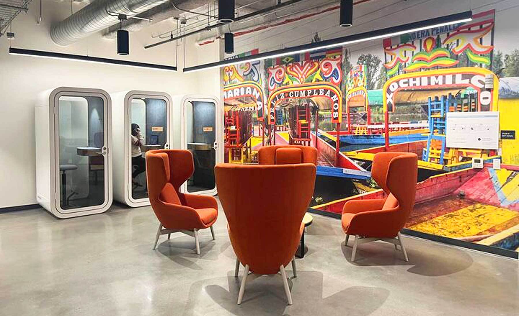 An image of a room with bright orange chairs in a circle. There is a colorful mural on the wall and phone call booths along another wall.