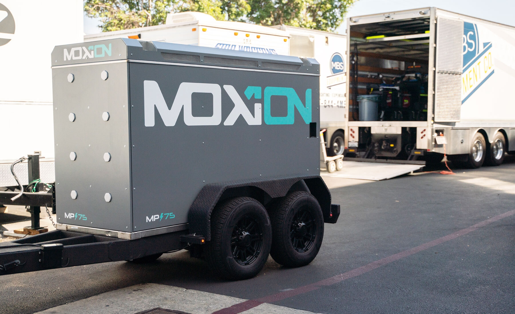 A small, gray trailer with the Moxion Power logo, situated in a parking lot in front of other larger trailers