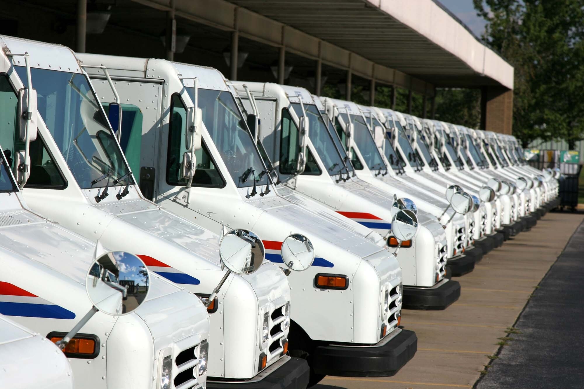 More than a dozen United States Postal Services delivery vehicles lined up under the canopy of a Postal Service building.