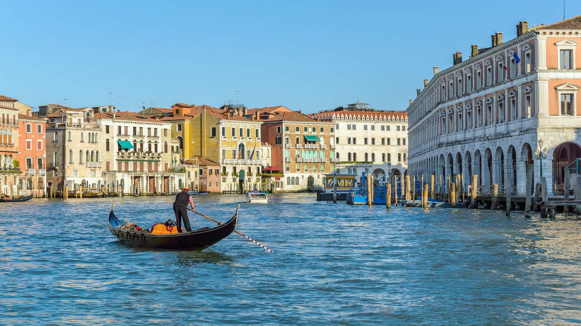 An image showing a large body of water with a person standing in a boat while rowing it. Buildings from an Italian city surround the water.