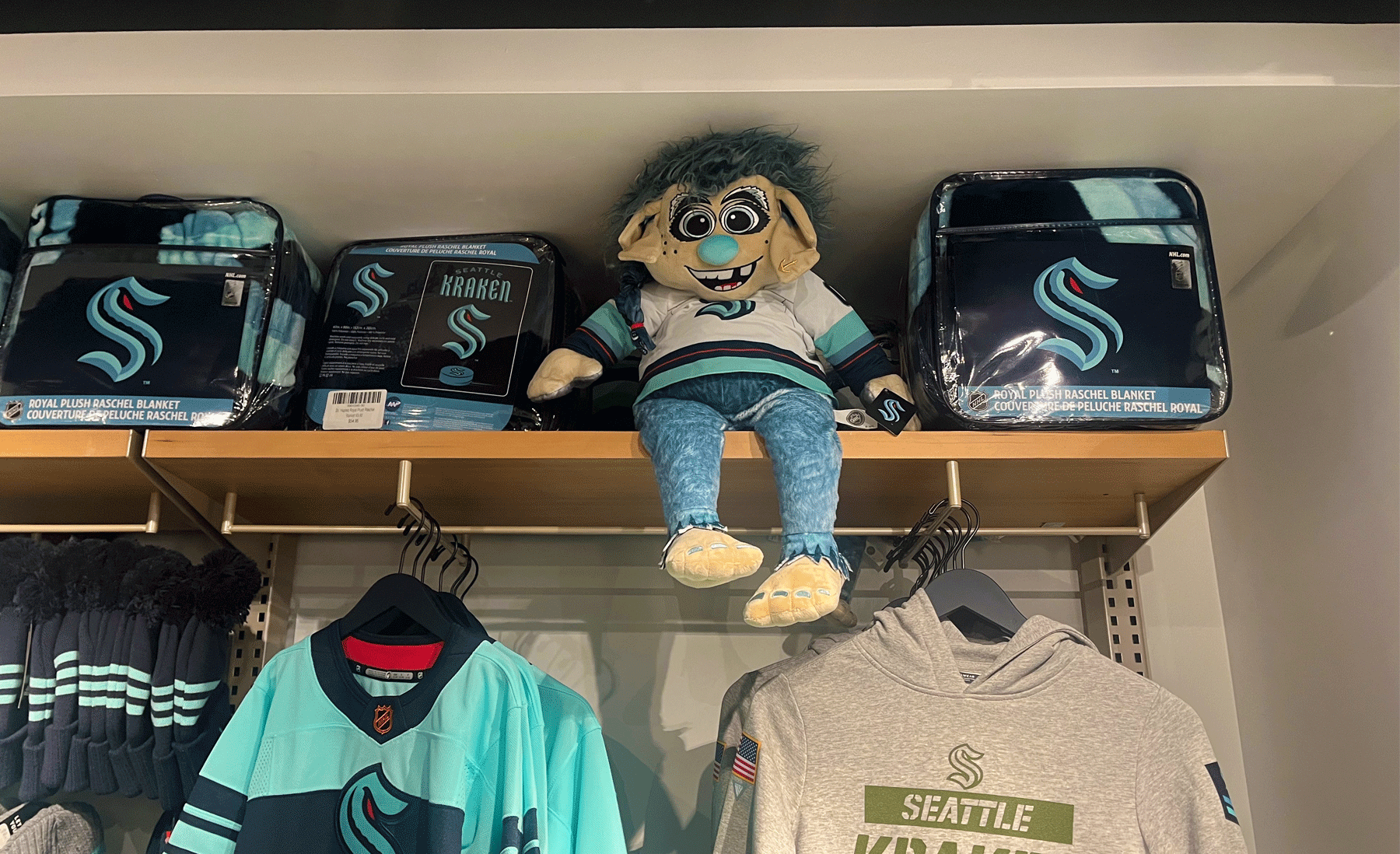 An image of a shelf of merchandise in the Seattle Kraken store. There is a plush toy of the troll mascot, Buoy sitting on the shelf in a Seattle Kraken jersey. 