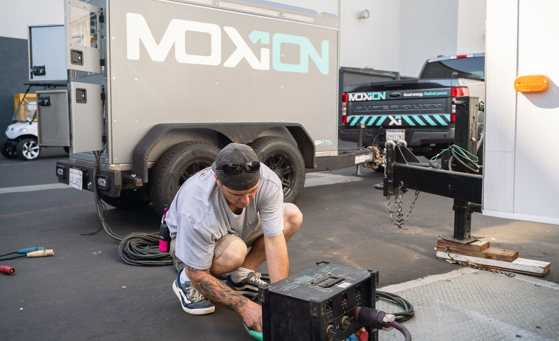 Man in casual clothes bending down to adjust some technical equipment in the backlot of a movie studio. Behind him is a Moxion truck and trailer.