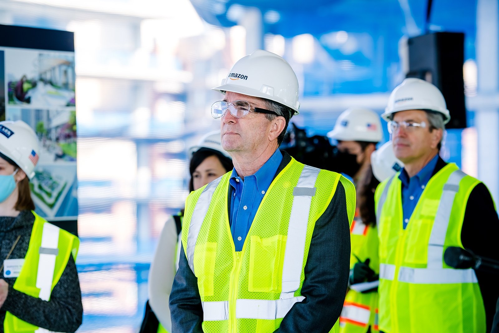 An image of a man wearing a hard hat that says "Amazon" and a yellow safety vest looking and listening to someone off camera while standing at a construction site. The man is accompanied by a group of people with similar safety gear on.
