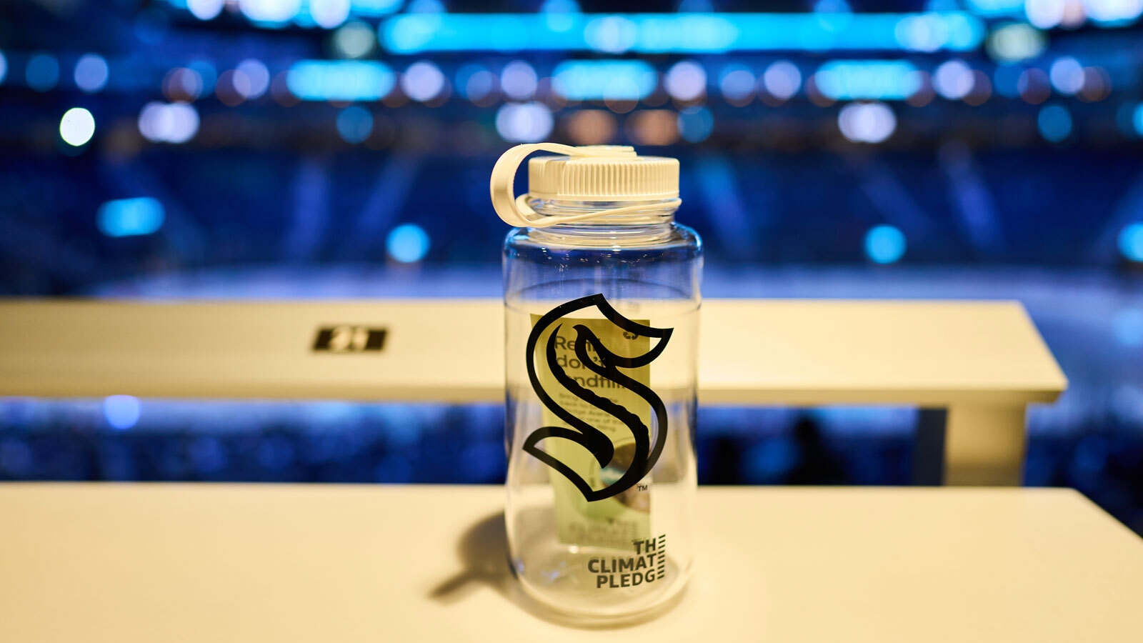An image of a clear water bottle with the Seattle Kraken logo on it. It's sitting on top of a table with the Climate Pledge Arena in the background.